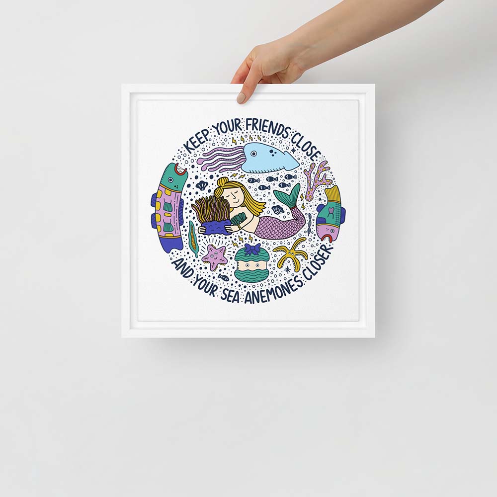 Hand holding artwork of mermaid hugging sea anemone surrounded by colorful ocean creatures with the phrase “Keep your friends close and your sea anemones closer” – framed square canvas wall art