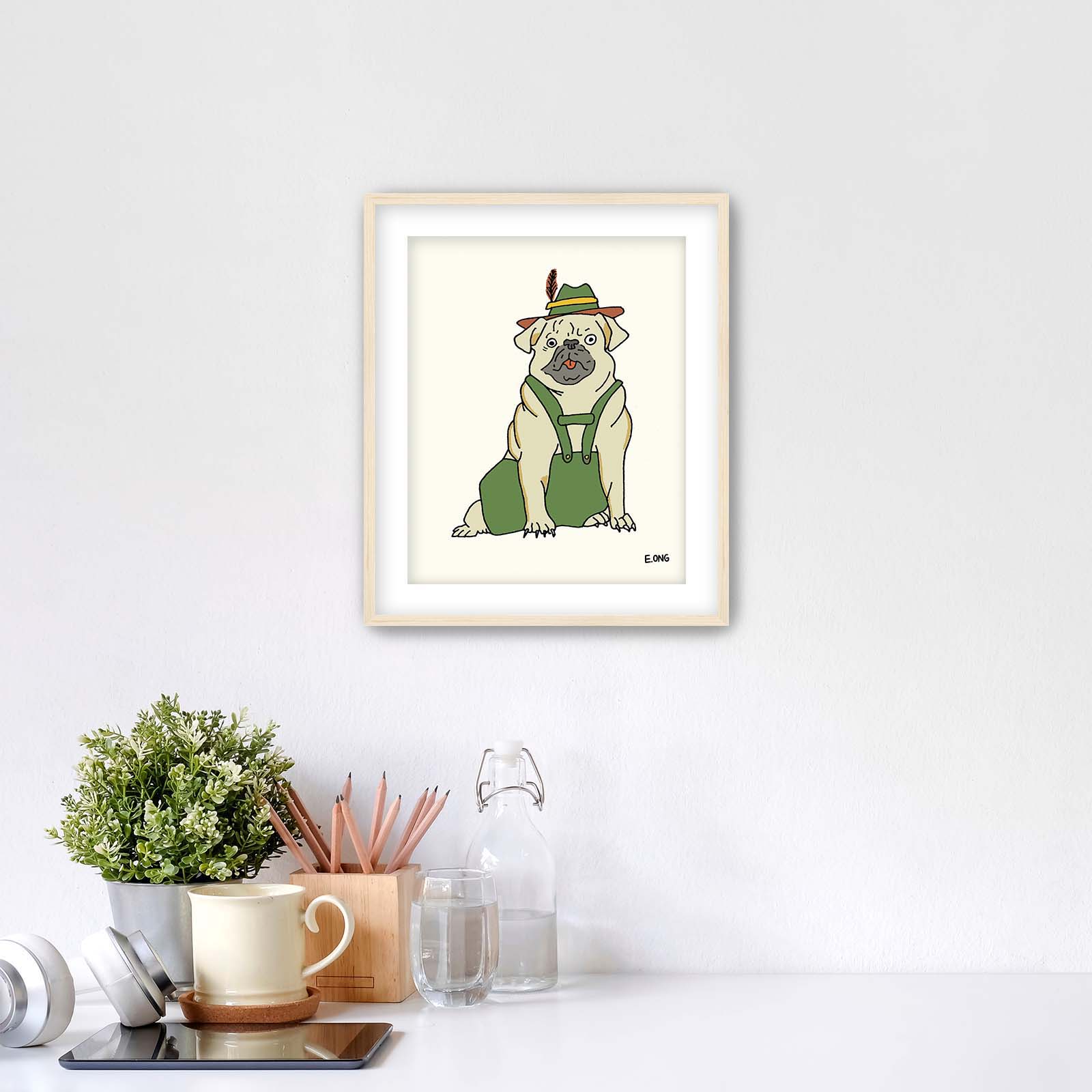 Whimsical pug in lederhosen art print by Erwin Ong shown in a light maple frame, styled above a clean white desk with cozy decor – fun wall decor for dog lovers and kids' rooms.