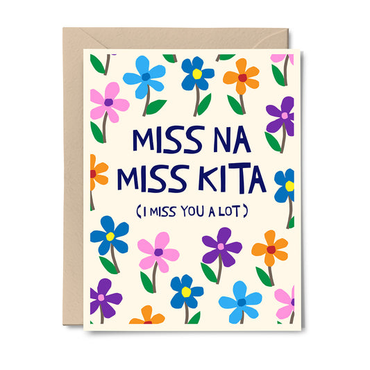 A square greeting card with a kraft envelope behind it, featuring the text 'MISS NA MISS KITA (I MISS YOU A LOT)' in dark blue lettering, surrounded by colorful illustrated flowers on a cream background.