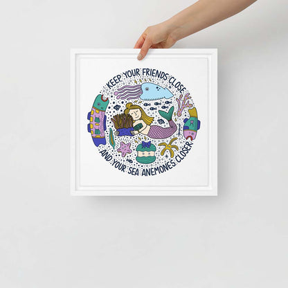 Hand holding artwork of mermaid hugging sea anemone surrounded by colorful ocean creatures with the phrase “Keep your friends close and your sea anemones closer” – framed square canvas wall art
